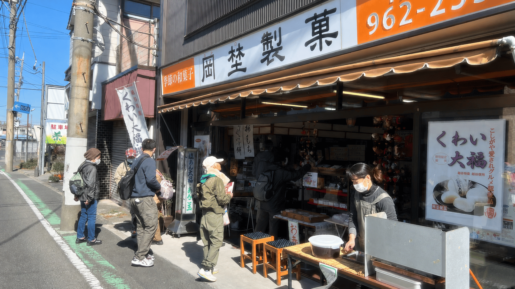 岡埜製菓店の店先。「越谷特産くわい大福」ののぼりが立ち、日よけテントの下にお客さまが並ぶ